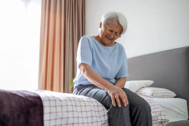 Tired woman in her 50s sitting on the edge of a bed, holding her knee in discomfort